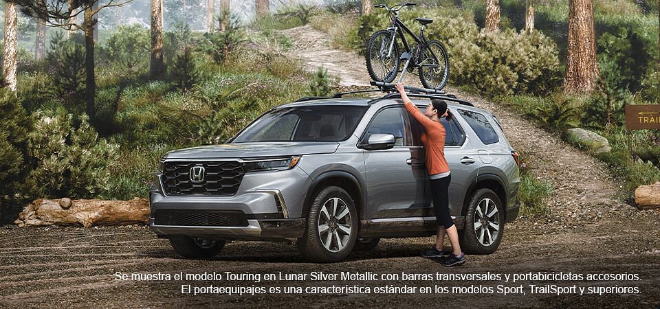 Mujer colocando una bicicleta en el Honda Pilot 2023 en color Lunar Silver metalizado con barras transversales accesorias y enganche para bicicleta en un aparcamiento forestal.